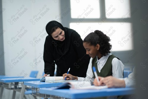 A Saudi teacher, who interacts with the students while explaining the lesson, in the classroom, tables and school supplies appear