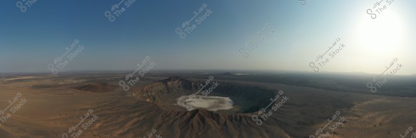 Aerial view of a massive crater in an open desert landscape. The crater is prominently visible amidst the sandy terrain, contrasting with the darker surrounding ground. The sky is clear blue with a bright sun on the horizon, imparting a sense of tranquility and vastness.