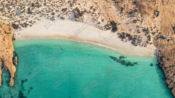 Aerial view of a sandy beach bordered by clear turquoise sea waters. The beach is surrounded by rocky terrain and scattered vegetation on the sand.