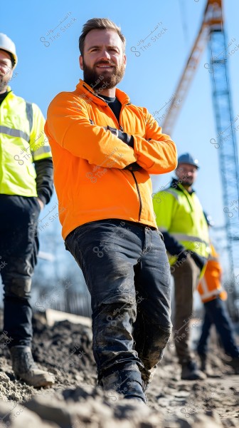 The image shows a group of workers at a construction site. The main person in the image stands confidently, wearing an orange jacket and dirt-stained dark pants, suggesting leadership or supervision. In the background, other workers in yellow safety jackets and helmets are visible, with a construction crane set against a clear blue sky. The ground around them is covered in mud and dirt.