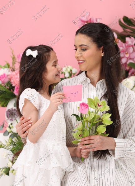 An image of a woman and a girl smiling at each other against a background of various flowers. The woman is wearing a striped black and white shirt and holding a bouquet of green flowers and pink roses. The girl is wearing a white dress with a white hair accessory and is holding a pink card with \"I love you, Mom\" written on it along with a small heart.