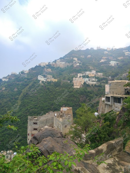 An image showing a series of white buildings constructed on lush green mountains. The buildings are spread along the slopes in a tiered manner, surrounded by dense greenery. In the foreground, there are some trees and large rocks, with a cloudy sky above.