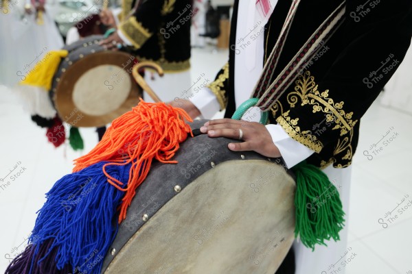 Image of a cultural performance showing a person in traditional attire decorated with golden embroidery. The individual is holding a musical instrument resembling a drum, covered with colorful threads in blue and orange. The embroidered sleeves and traditional clothing suggest cultural heritage.