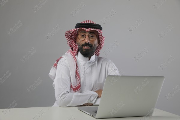 A portrait of a man wearing traditional Saudi attire with a red and white checkered headdress and black agal, seated in front of a laptop on a white desk, against a gray background. He is smiling and wearing glasses.
