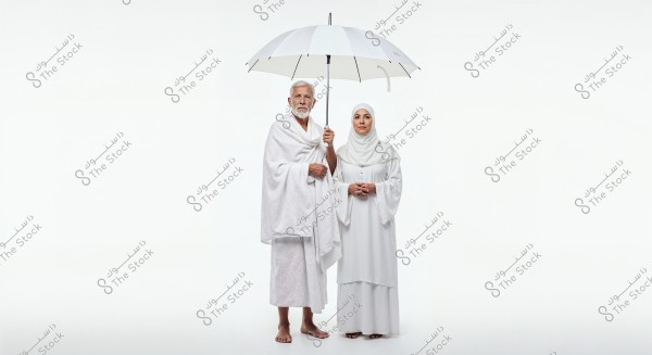 Image of a man and woman dressed in white Ihram clothing standing under a white umbrella. The man is holding the umbrella and is wearing traditional Ihram attire, while the woman is wearing a hijab and a white dress. They appear to be preparing for Hajj or Umrah. The background is simple and white.