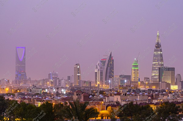 Nighttime view of Riyadh\'s skyline in Saudi Arabia, showcasing prominent buildings like the Kingdom Centre and Al Faisaliah Tower with various illuminated lights, with traditional houses in the foreground under a pinkish sky.