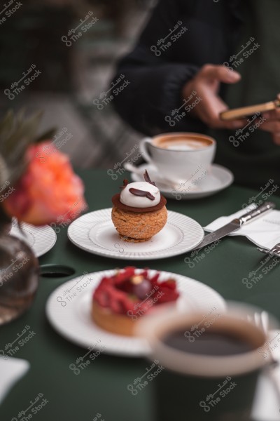 A person sitting at a table using their phone, with a cup of coffee and a dessert plate topped with chocolate and cream, and a blurred background of a flower and some plates.