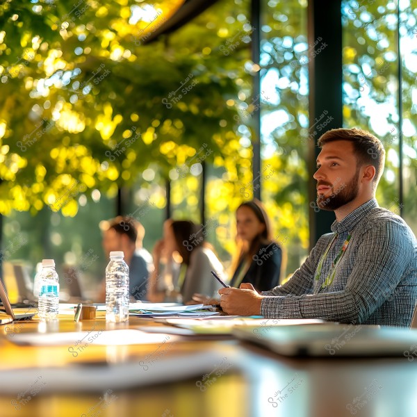 A man sits in a brightly lit meeting room adorned with sunlit green plants. He is wearing a long-sleeved striped shirt and holding a pen while focusing on a presentation. In the background, other people are seated at a table with papers and water bottles scattered around. The background walls are glass, allowing natural light to illuminate the plants outside.
