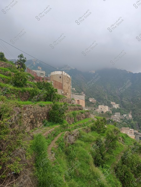 The image shows a natural landscape in a foggy mountainous region. Terraces covered with greenery cascade down the mountain, with traditional houses scattered along the slope. The houses are designed in a traditional architectural style with stone walls and flat roofs. This area is likely associated with the Sarawat Mountains in Saudi Arabia, evidenced by the architecture and surrounding vegetation.
