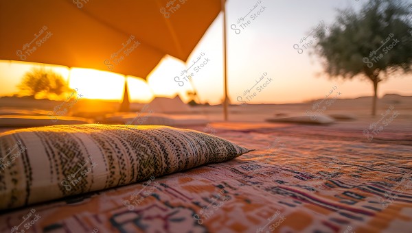 A sunset scene in the desert featuring a traditional tent in the foreground, with a woven cushion on the patterned ground. The sun is setting with golden hues, and there is a tree on the right side in the background. The atmosphere is calm and warm, reflecting the Bedouin culture in a desert environment.