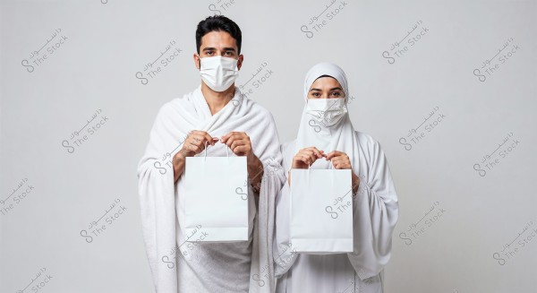 An image of a man and a woman wearing white Ihram clothing and face masks. The man is holding a white bag and is dressed in the traditional unstitched Ihram garment, while the woman is fully covered with a hijab and also holding a white bag. The background is simple and neutral-colored.