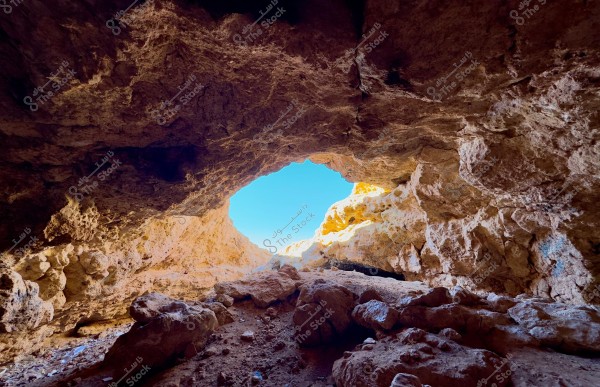 The image shows a cave with a large opening overlooking a bright blue sky. The cave\'s interior walls are made of natural light brown rocks of varying sizes and shapes, covered with some shadows. The cave floor is scattered with rocks and dirt.
