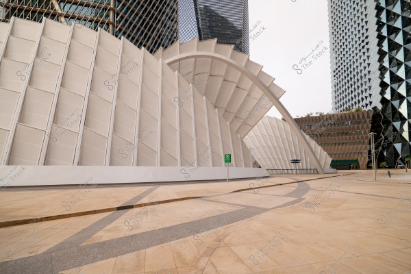 Image of a modern building with a unique architectural design, featuring slanted white walls and a large arch. The building is located in an urban area with distinctive glass towers in the background. The ground has a geometric pattern complementing the architectural style of the surrounding buildings.