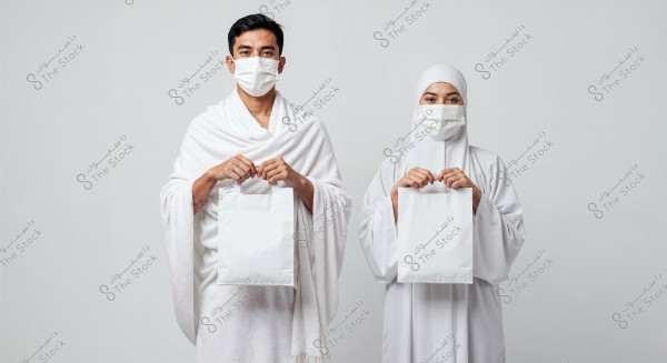 A portrait of a man and woman wearing white Ihram clothing and white medical masks, standing side by side and holding white paper bags. The background is a neutral light color.