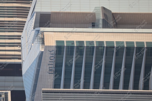 Image of a modern multi-story building with a glass facade and sharp architectural lines. The word \"Hotel\" is prominently displayed on the side of the building, which appears to be part of a larger complex with a similar architectural structure in the background. Focus is on linear shapes and contemporary design.