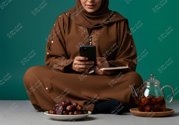 Image of a woman sitting on the floor in traditional brown embroidered clothing, holding a mobile phone and a small notebook. In the foreground, there is a plate of dates and a glass teapot on a wooden tray. The background is dark green.
