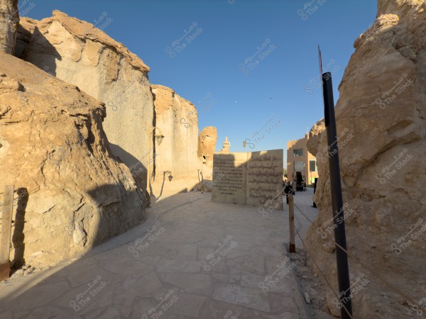 ممر مرصوف محاط بتكوينات صخرية طبيعية في البيئة الصحراوية, تظهر واجهات مبانٍ تقليدية على مسافة. هناك لوحة كبيرة مغطاة بنقوش وأعمدة إنارة على طول الممر. السماء زرقاء وصافية في الخلفية.