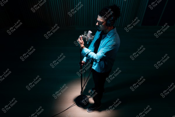 شخص يرتدي سماعات رأس ويقف أمام ميكروفون في استوديو تسجيل مظلم.