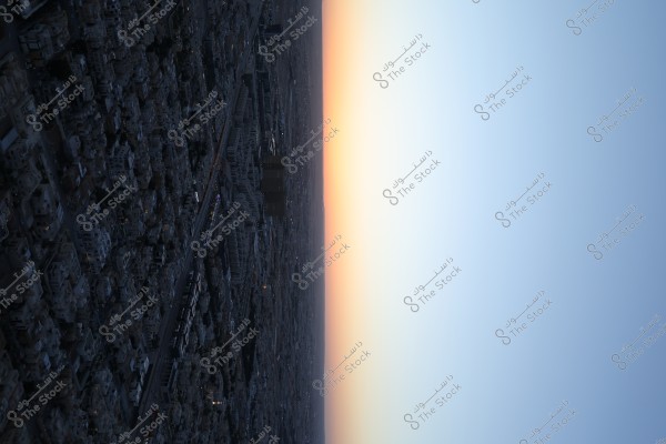 An image of a cityscape taken from a high elevation during dawn or dusk. The photo shows the city with various buildings and streets, while the sky displays a gradient of colors from light blue to orange, indicating sunrise or sunset.