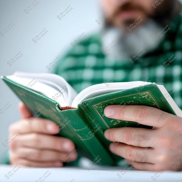 Image of a man with a gray beard wearing a green plaid shirt, holding and reading a book with a decorative green cover. The focus is on the hands and the book, while part of his face is blurred in the background.