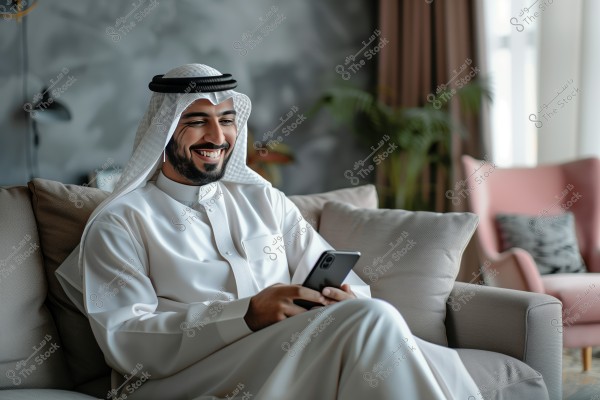 A man is sitting and smiling on a sofa while holding a smartphone. He is wearing a white traditional robe and head cover. The room is decorated with plants and modern decor, and there is a pink chair in the background.