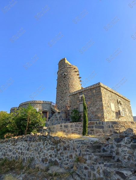 صورة لمبنى حجري قديم مكون من طابقين في يوم صافٍ ومشمس, حيث يظهر المبنى بجدران حجرية وبرج دائري طويل. يحيط بالمبنى بعض النباتات والأشجار الخضراء, ويظهر على يسار المشهد درج حجري يؤدي إلى المدخل. السماء زرقاء وصافية, مما يعزز جمال المشهد.