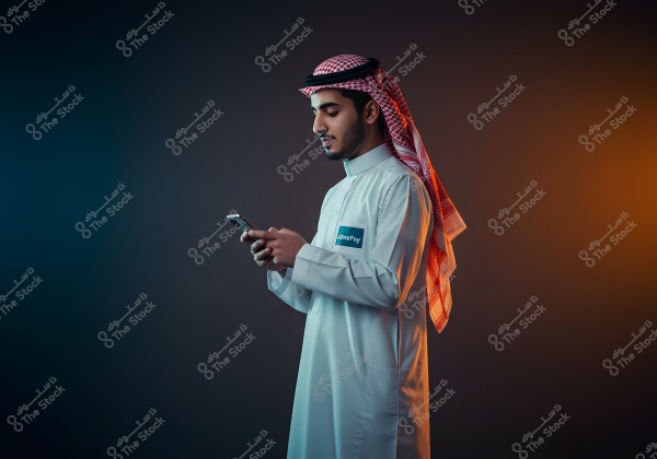Portrait of a man wearing traditional Gulf attire, including a white thobe and a red and white checkered ghutra, standing against a gradient background of orange and blue hues. The person is holding a mobile phone, focused on the screen.