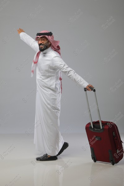 Image of a man wearing traditional Saudi attire, consisting of a white thobe and a red and white ghutra with a black agal. The man is smiling broadly and stands in a playful pose, extending his arm forward as if pointing in the direction he\'s heading. He is holding a red wheeled suitcase. He appears to be preparing for a journey or new adventure. The background is a solid gray color.
