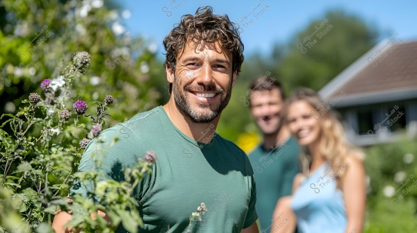 An image of a smiling man wearing a green shirt standing in a lush garden filled with plants and flowers. In the background, a house is visible with sunlight illuminating the scene, and two other smiling people are out of focus.