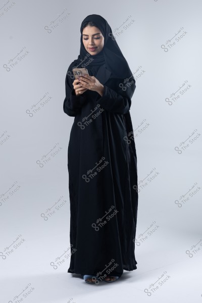 A portrait of a woman wearing a black abaya and a black hijab, standing while using a smartphone. The background is white, and the lighting is soft, highlighting her calm demeanor. The clothing suggests she is likely from the Gulf region.