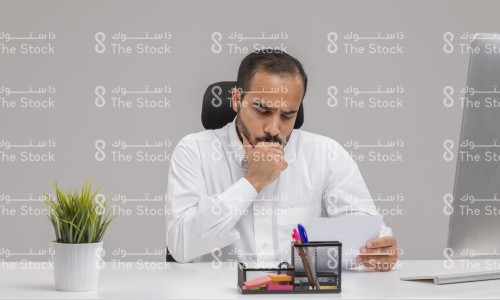 شاب سعودي يرتدي الثوب السعودي يتصفح الأوراق أثناء عمله في مكتبه