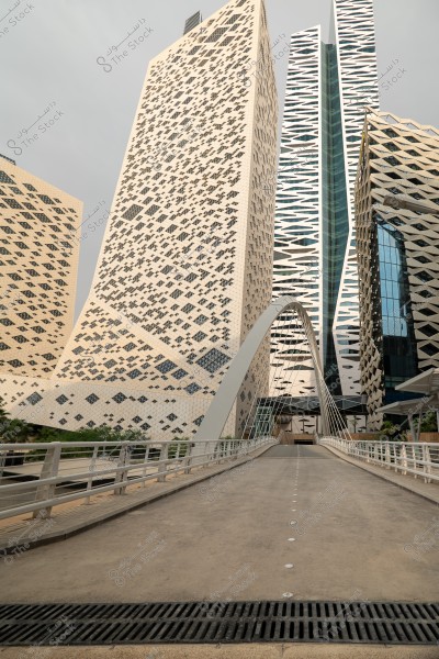 A modern pedestrian bridge leads to a group of tall architectural towers with intricate geometric designs in an urban area. The towers are covered with geometric patterns and glass structures, with a gray sky in the background.