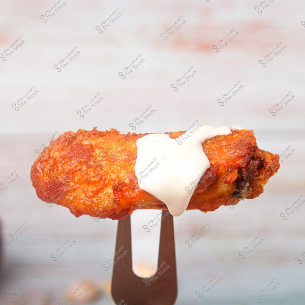 The image shows a piece of fried chicken wing topped with white sauce, placed on a fork against a blurred light-colored background.