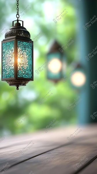 A traditional hanging lantern made of metal with intricate designs and turquoise glass, emitting a warm glow. Two similar blurred lanterns are seen in the background, with a brown wooden surface below. The background features an out-of-focus green outdoor setting.