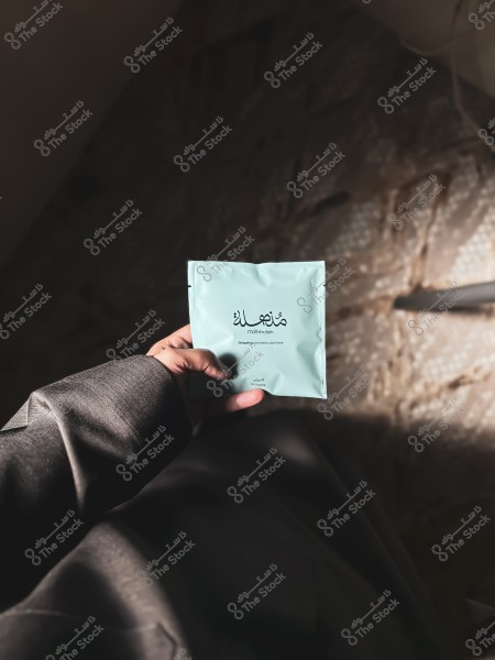 Image of a person's hand holding a small light blue packet with the text "مذهلة" in Arabic and "mohtheleh" in English. The hand is wearing a dark traditional garment, suggesting traditional attire. The background is blurred and dimly lit, adding a natural and calm atmosphere to the image.