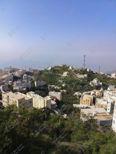 منظر طبيعي لمدينة تقع في منطقة جبلية, تضم منازل ومباني متعددة الألوان محاطة بالأشجار والمساحات الخضراء. هناك أبراج اتصالات ظاهرة في الخلفية تحت سماء زرقاء صافية.