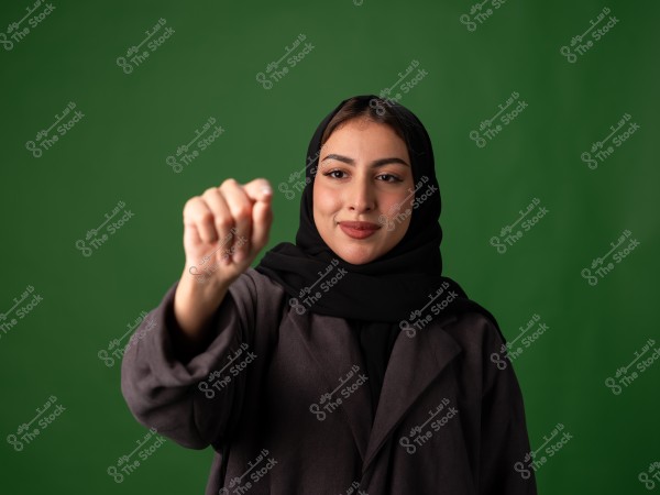 صورة لامرأة تقف أمام خلفية خضراء. ترتدي عباءة سوداء وحجابًا أسود, وتظهر وهي تمد يدها قليلاً للأمام. تعابير وجهها تبدو مبتسمة قليلاً.
