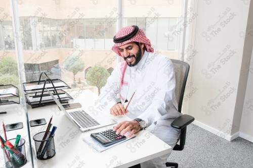 رجل سعودي يرتدي الزي السعودي التقليدي يعمل في مكتب بواجهة زجاجية ويستخدم قلم رصاص لتدوين الملاحظات