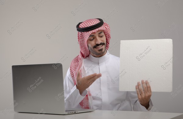 A man wearing traditional Saudi attire with a red shemagh is sitting at a desk in front of a laptop, smiling and gesturing towards a white board he is holding. The background is neutral and simple.