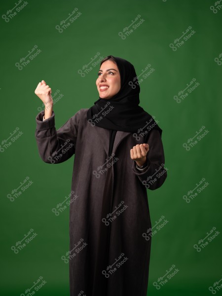 A portrait of a woman wearing a black abaya and a black hijab, standing and smiling with her hands raised in a gesture of joy or victory. The background is green.