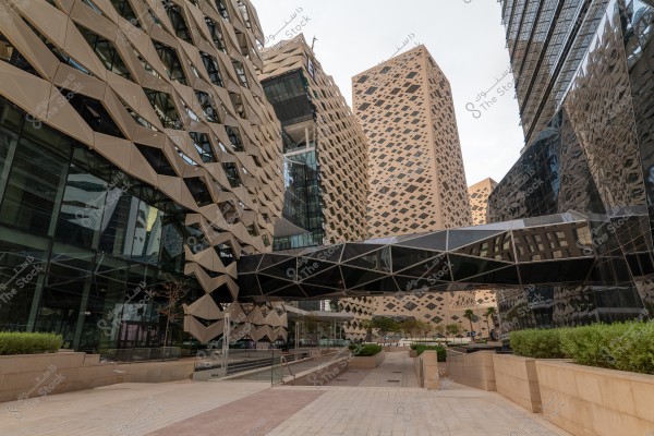 The image shows a group of modern buildings in an urban area. The buildings feature a unique architectural design with interlocking geometric patterns. In the center, there is a glass walkway connecting the buildings, and the scene is surrounded by wide pavements and some greenery. The sky is clear, with the view extending vertically upwards.