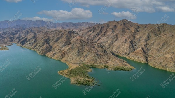 A scenic view of a brown mountain surrounded by a bluish-green river under a blue sky with white clouds. Green vegetation is seen growing along the riverbanks, adding a beautiful contrast to the barren mountain terrain. The mountains extend into the horizon, forming striking natural gradients.