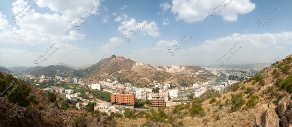 A panoramic view of a landscape showing a city surrounded by green hills and mountains. There are buildings of various sizes and colors with a road passing through them. The sky appears blue with scattered clouds in the background.