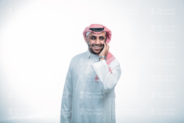 بورتريه رجل سعودي مبتسم يرتدي الزي السعودي التقليدي يتحدث عبر الجوال ، تعابير وجه تدل على السعادة ، حوار ممتع مع شخص ما