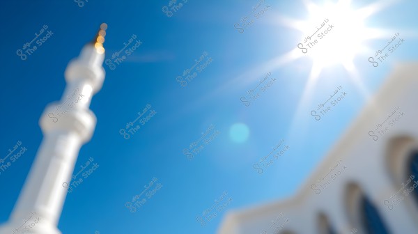 The image depicts a scene of bright sunlight in a blue sky, with a blurred silhouette of a white minaret on the left and a building with arches below. The details are out of focus, giving the scene an artistic touch.