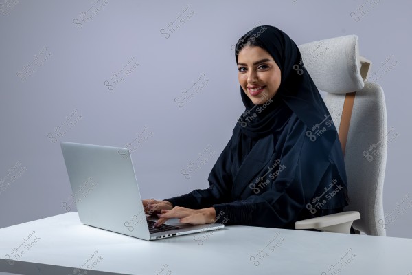 An image of a woman wearing a hijab and a black abaya, sitting on a modern office chair and working on a silver laptop on a white table. The background is light gray, giving the photo a professional and minimalist feel.