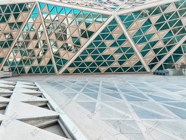 Image of a modern building facade with a unique geometric design, featuring three-dimensional windows in light blue and beige. The ground in front of the building has an interlocking square pattern. On the right corner, there are some seats and potted plants.