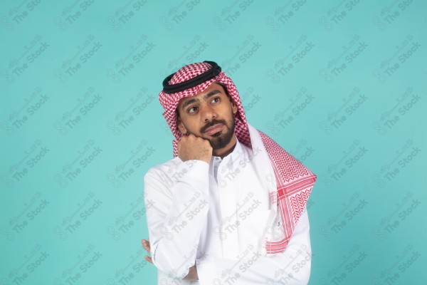 بورتريه رجل سعودي يرتدي الثوب يقف أمام خلفية تركوازية ، تعابير وجه تدل على التساؤل والحيرة والتفكير ، لحظة اتخاذ قرار ، صورة شخصية