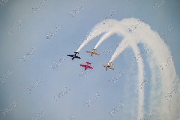 Four small airplanes flying in formation in the blue sky, emitting thick white smoke that arches behind them in organized trails. One aircraft is red while the others are black and two are light-colored.