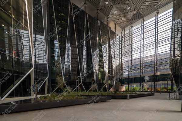 Image of a facade of a modern building made of glass and metal beams. The architectural design includes reflective glass that mirrors the surrounding environment, with interior lights adding a contemporary touch. There is a planted area at the bottom that adds a natural element to the industrial design, with a clean concrete floor and open spaces around it.